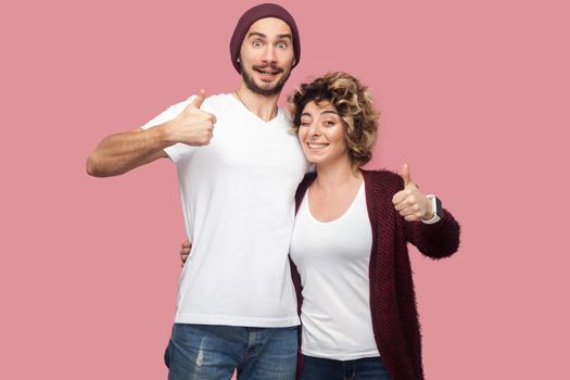 Awesome. Portrait of positive couple of friends in casual style standing, hugging and showing thumbs up with toothy smile, looking at camera. Isolated, indoor, studio shot, pink background