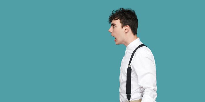 Profile side view portrait of crazy or shocked handsome hipster curly young businessman in classic casual white shirt and suspender standing, screaming. indoor studio shot isolated on blue background.
