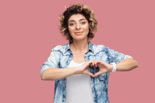 Portrait of happy young woman with curly hairstyle in casual blue shirt standing with heart hands shape gesture and looking at camera smiling. indoor studio shot, isolated on pink background.