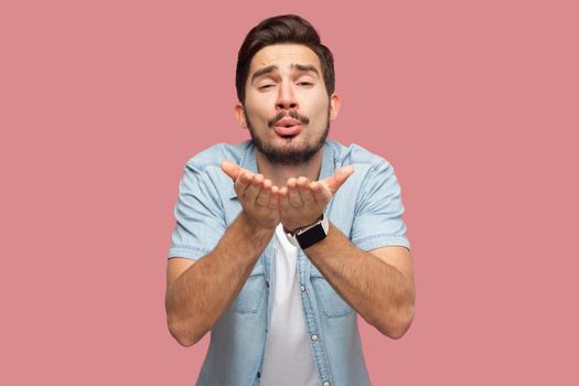 Portrait of happy in love handsome bearded young man in blue casual style shirt standing and looking at camera and sending air kiss. indoor studio shot, isolated on pink background.