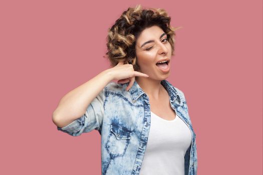 Call me. Portrait of funny young woman with curly hairstyle in casual blue shirt standing with call gesture and looking at camera with funny face. indoor studio shot, isolated on pink background.