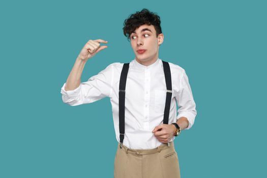 I need more. man showing a little bit gesture with finger. Portrait of handsome hipster curly young businessman in classic casual white shirt and suspender. studio shot isolated on blue background.