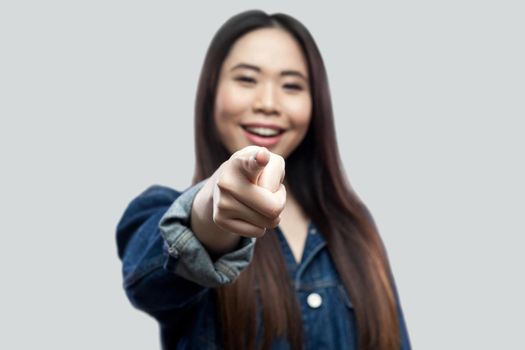 Portrait of excited beautiful brunette asian young woman in casual blue denim jacket with makeup standing toothy smiling and pointing at camera. indoor studio shot, isolated on light grey background.