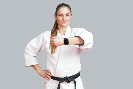 Beautiful satisfied proud young blonde athletic woman posing and showing her smart watch, smile and looking at camera. Woman using fitness bracelet. Indoor studio shot, isolated on grey background