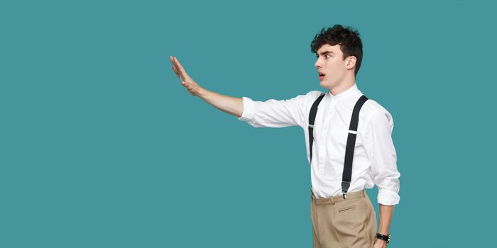 Profile side view portrait of serious handsome hipster curly young businessman in classic casual white shirt and suspender standing with stop sign gesture. studio shot isolated on blue background.