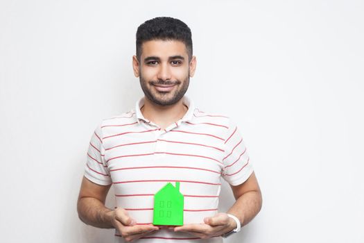 Portrait of cheerful positive glad optimistic young adult man in t-shirt have dream and holding paper small house, planning new home to buy. Indoor, isolated, studio shot, copy space, white background