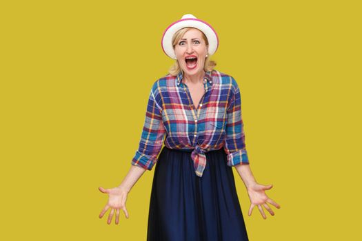 Portrait of nervous aggressive stylish mature woman in casual style with hat standing, raised arms with angry face and looking at camera screaming. indoor studio shot isolated on yellow background.