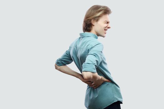 Kidney or spine pain. Profile side view portrait of handsome long haired blonde young man in blue casual shirt standing touching his painful back. indoor studio shot, isolated on light grey background