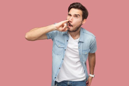 Portrait of crazy handsome bearded young man in blue casual style shirt standing and drilling his nose with funny face. indoor studio shot, isolated on pink background.
