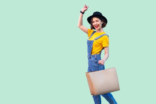 Cheerful beautiful young girl in hipster wear in denim overalls and black hat holding shop bag toothy smile, celebrating great shopping with raised arm, looking at camera. Studio shot,green background