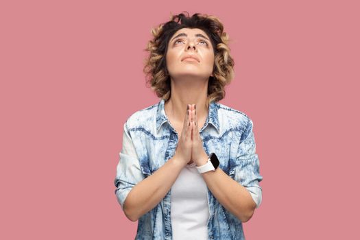 My god please help me. Portrait of hopeful young woman with curly hairstyle in casual blue shirt standing with palm hand and praying or begging. indoor studio shot, isolated on pink background.