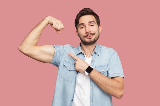 I am strong and I can do anything. Portrait of proud handsome bearded young man in blue casual shirt standing pointing and showing his strong bicep. indoor studio shot, isolated on pink background.