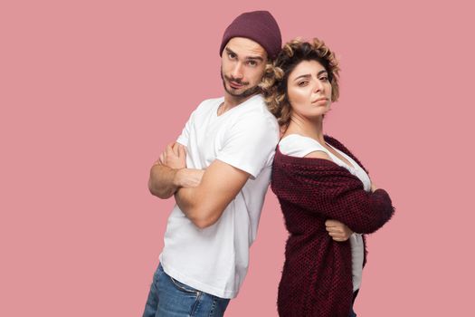 Portrait of serious couple of friends in casual style standing back to back, crossing arms and looking at camera with proud face. Isolated, indoor, studio shot, pink background