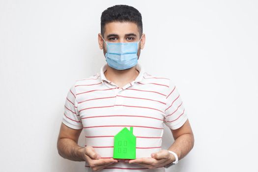 Stay at home, stay safe. Positive man with surgical medical mask holding green small house, looking at camera kindly, reassuring face on quarantine time. Indoor isolated, studio shot, white background