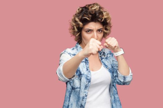 Portrait of serious young woman with curly hairstyle in casual blue shirt standing with boxing fists, looking at camera and ready to attack. indoor studio shot, isolated on pink background.