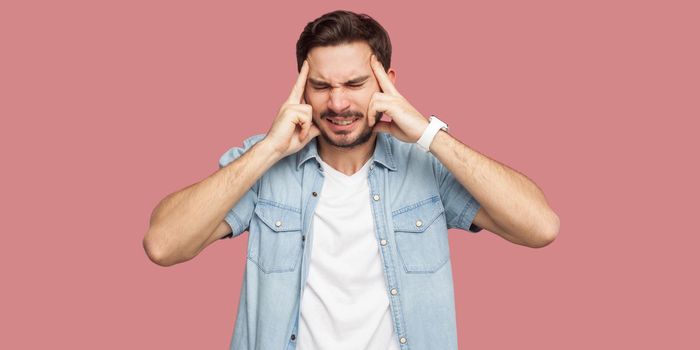 Headache, confusion or thinking. Portrait of sad handsome bearded young man in blue casual shirt standing and holding his head, thinking or endure. indoor studio shot, isolated on pink background.