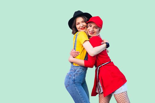 Couple of beautiful stilysh hipster best friends in fashionable clothes standing, hugging with love, glad to see each other, looking at camera. Indoor studio shot, isolated on green background