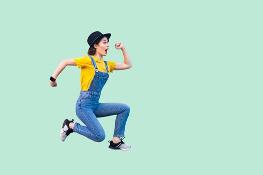 Profile side view portrait of surprised funny young hipster girl in blue denim overalls, yellow shirt and black hat jumping in super mario style. indoor studio shot isolated on light green background.
