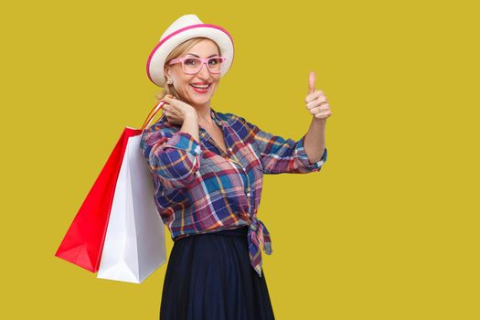Happy modern aged woman in white hat and in checkered shirt standing, holding shopping bags and showing thumbs up with toothy smile, looking at camera. Indoor, studio shot, yellow background, isolated