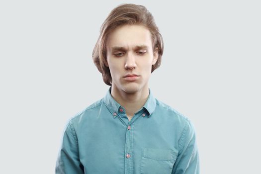 closeup portrait of sad worried handsome long haired blonde young man in blue casual shirt standing, holding head down and thinking what to do. indoor studio shot, isolated on light grey background.