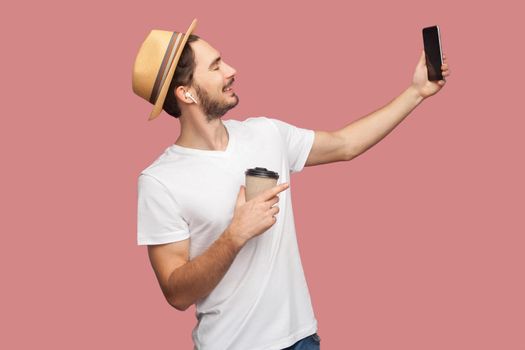 Side view portrait of handsome bearded young hipster man in white shirt and casual hat posing and making selfie photo with smile, blogger lifestyle. Indoor,isolated, studio shot, pink background