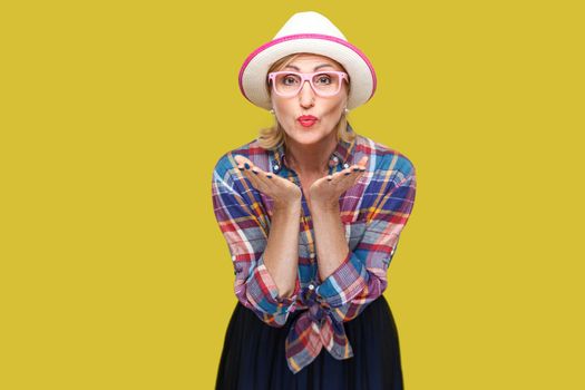Portrait of funny modern stylish mature woman in casual style with hat and eyeglasses standing sending kiss and looking at camera with romantic expression. studio shot isolated on yellow background.