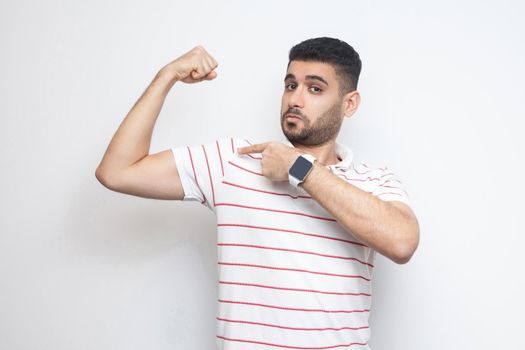 I am strong. Portrait of proud handsome bearded young man in striped t-shirt standing, pointing at his bicep and looking at camera with satisfied face. indoor studio shot, isolated on white background