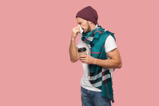 Portrait of sickness modern bearded young hipster man in white shirt and casual hat standing,sneezes of napkin, holding hot drink in paper cup. Indoor, isolated, studio shot, pink background