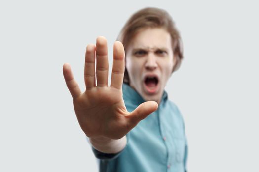 Stop it. Portrait of angry handsome long haired blonde man in blue shirt standing with stop hand gesture sign and looking at camera screaming. indoor studio shot, isolated on light grey background.