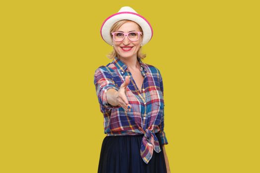 Portrait of happy modern stylish mature woman in casual style with hat and eyeglasses standing with toothy smile, looking and giving hand to greeting. indoor studio shot isolated on yellow background.