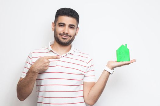 Want you own home? Happy young realtor man in t-shirt holding green paper small house and propose you to planning new home to buy. Indoor, isolated, studio shot, copy space, white background