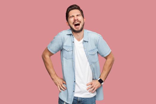 Portrait of funny careless handsome bearded young man in blue casual style shirt standing with hands on waist and laughing with closed eyes. indoor studio shot, isolated on pink background.