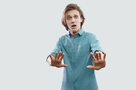 Portrait of scared or shocked handsome long haired blonde young man in blue casual shirt standing with blocking hand and looking at camera. indoor studio shot, isolated on light grey background.