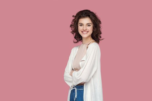 Portrait of satisfied beautiful brunette young woman with curly hairstyle in casual style standing and looking at camera with toothy smile and crossed arms. studio shot isolated on pink background.