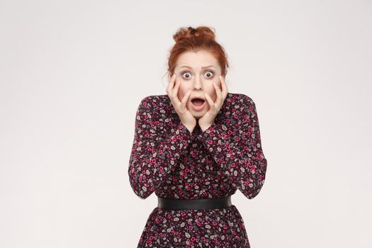 Human face expressions and emotions. Redhead woman screaming with shock, holding hands on her cheeks. Isolated studio shot on gray background.