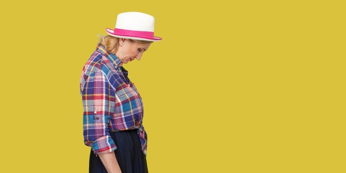 Profile side view portrait of sad alone stylish mature woman in casual style with hat standing, holding head down, frowning and crying. indoor studio shot isolated on yellow background.