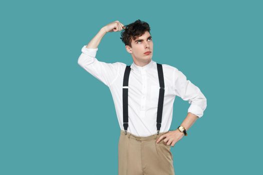 Confused man looking away and thinking. Portrait of handsome hipster curly young businessman in classic casual white shirt and suspender standing. indoor studio shot isolated on blue background.