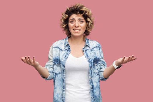 Portrait of confused or shocked young woman with curly hairstyle in casual blue shirt standing with raised arms and looking at camera worried. indoor studio shot, isolated on pink background.