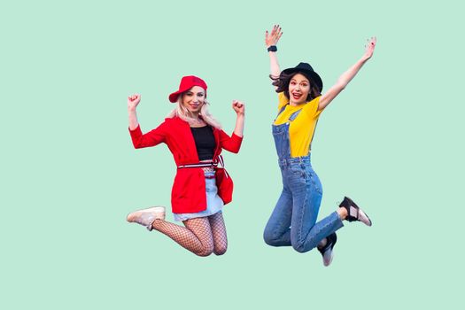 Body size portrait of two happy screaming stylish hipster girls in fashionable clothes are jumping up in the air and looking at camera with raised arms. Indoor studio shot,isolated on green background
