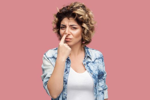 You are liar. Portrait of serious young woman with curly hairstyle in casual blue shirt standing with finger on her nose and showing lie gesture. indoor studio shot, isolated on pink background.
