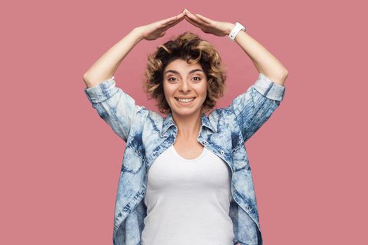 Portrait of happy young woman with curly hairstyle in casual blue shirt standing with roof gesture hands on head and looking at camera with toothy smile. indoor studio shot isolated on pink background