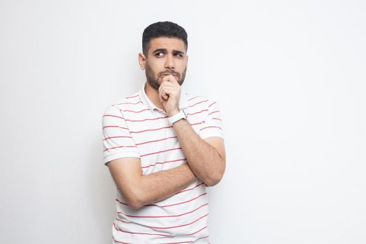 Portrait of thoughtful handsome bearded young man in striped t-shirt standing, looking away and thinking what to do. indoor studio shot, isolated on white background.