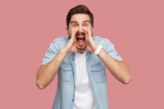 Portrait of crazy handsome bearded young man in blue casual style shirt standing with hand on face, looking at camera and screaming. indoor studio shot, isolated on pink background.