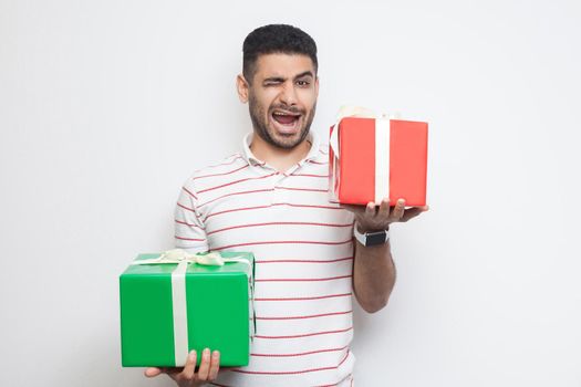 Portrait of positive attractive young adult man in t-shirt standing, holding two big gift box and winking, looking at camera. Studio shot, white background, isolated, indoor, copy space