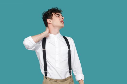 Neck pain. Man holding his painful or injury pain. Portrait of handsome curly young businessman in classic casual white shirt and suspender standing. indoor studio shot isolated on blue background.