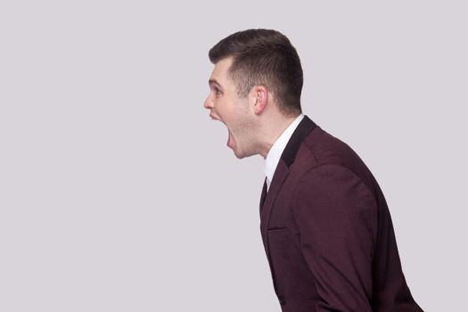 Profile side view portrait aggressive of handsome young man in violet suit and white shirt, standing and screaming with nervous face. indoor studio shot, isolated on grey background.