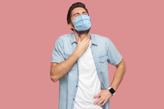 Throat pain or cold flu. Portrait of sad sick young man with surgical medical mask in blue shirt standing and holding his painful neck and screaming. indoor studio shot, isolated on pink background