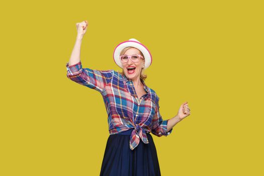 Portrait of happy surprised modern stylish mature woman in casual style with hat and eyeglasses standing and celebrating her victory and looking at camera. studio shot isolated on yellow background.