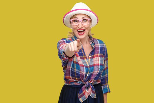 Portrait of funny surprised modern stylish mature woman in casual style with hat and eyeglasses standing, looking and pointing at camera with amazed face. studio shot isolated on yellow background.