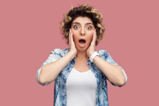 Portrait of shocked young woman with curly hairstyle in casual blue shirt standing with big eyes and open mouth and looking at camera surprised. indoor studio shot, isolated on pink background.